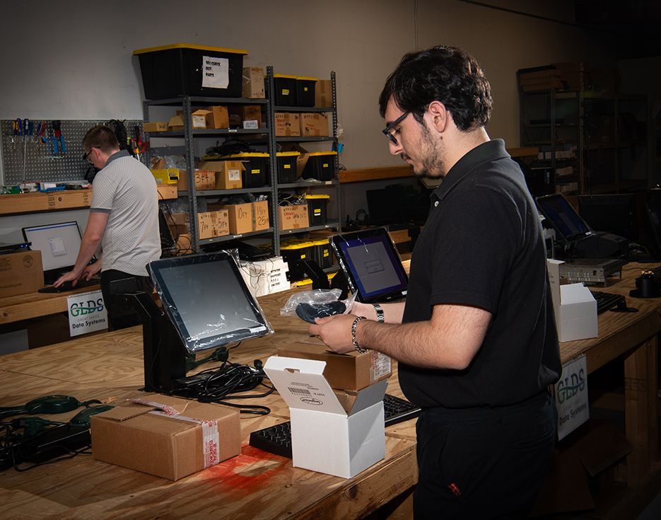Person packaging items at a warehouse workbench with laptops, boxes, and shelving in the background.