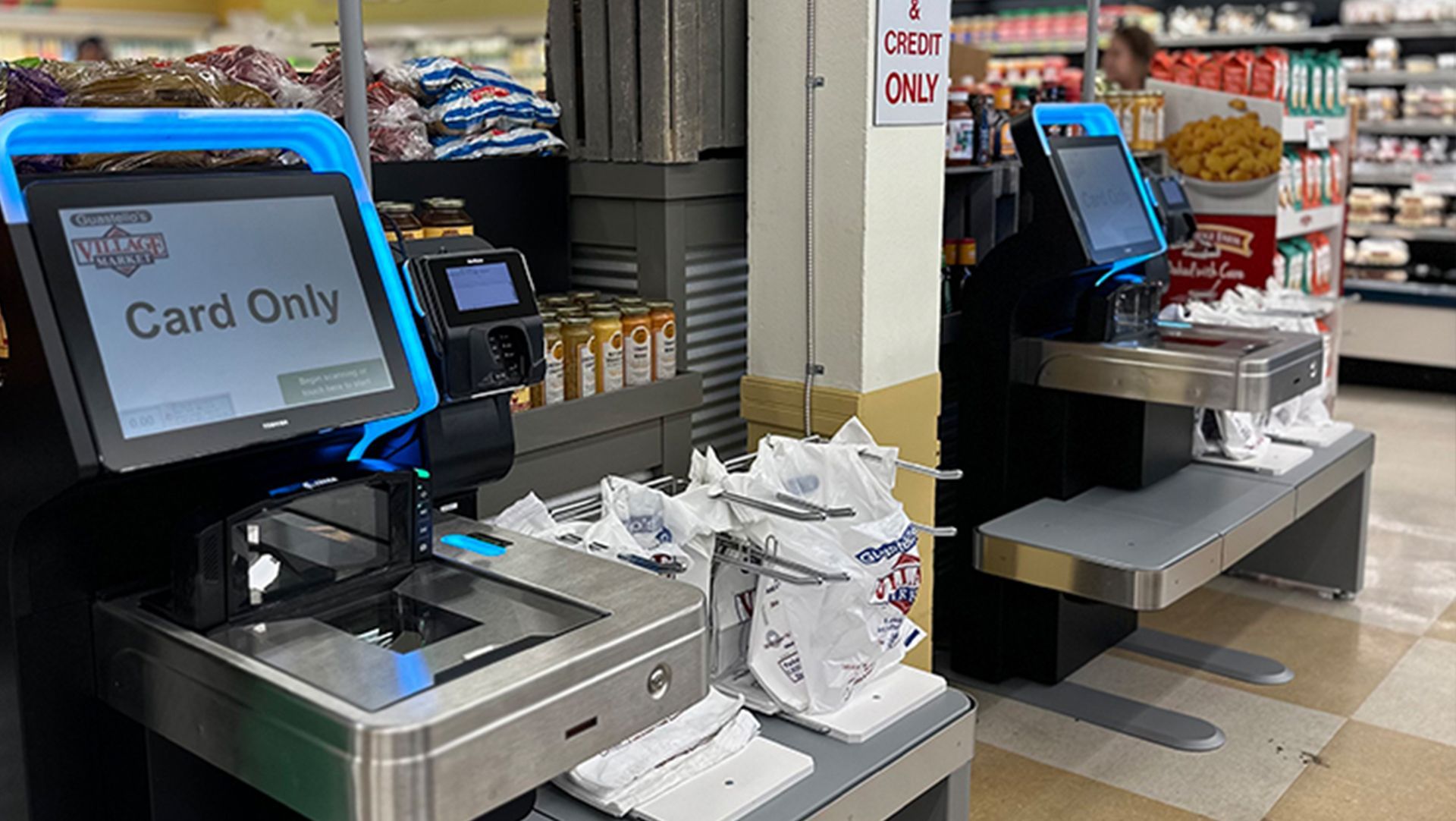 Two self-checkout kiosks in a store, one labeled “Card Only,” with bags and a shopping cart nearby.