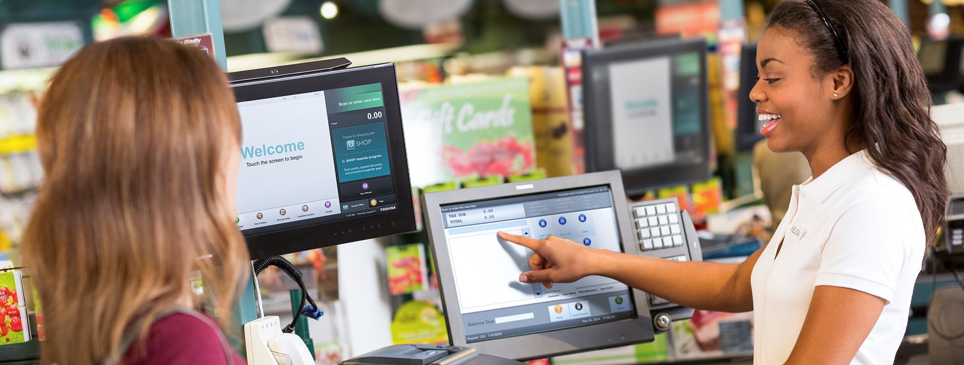 Two women shopping at a self-checkout kiosk, one tapping the touchscreen in a store.