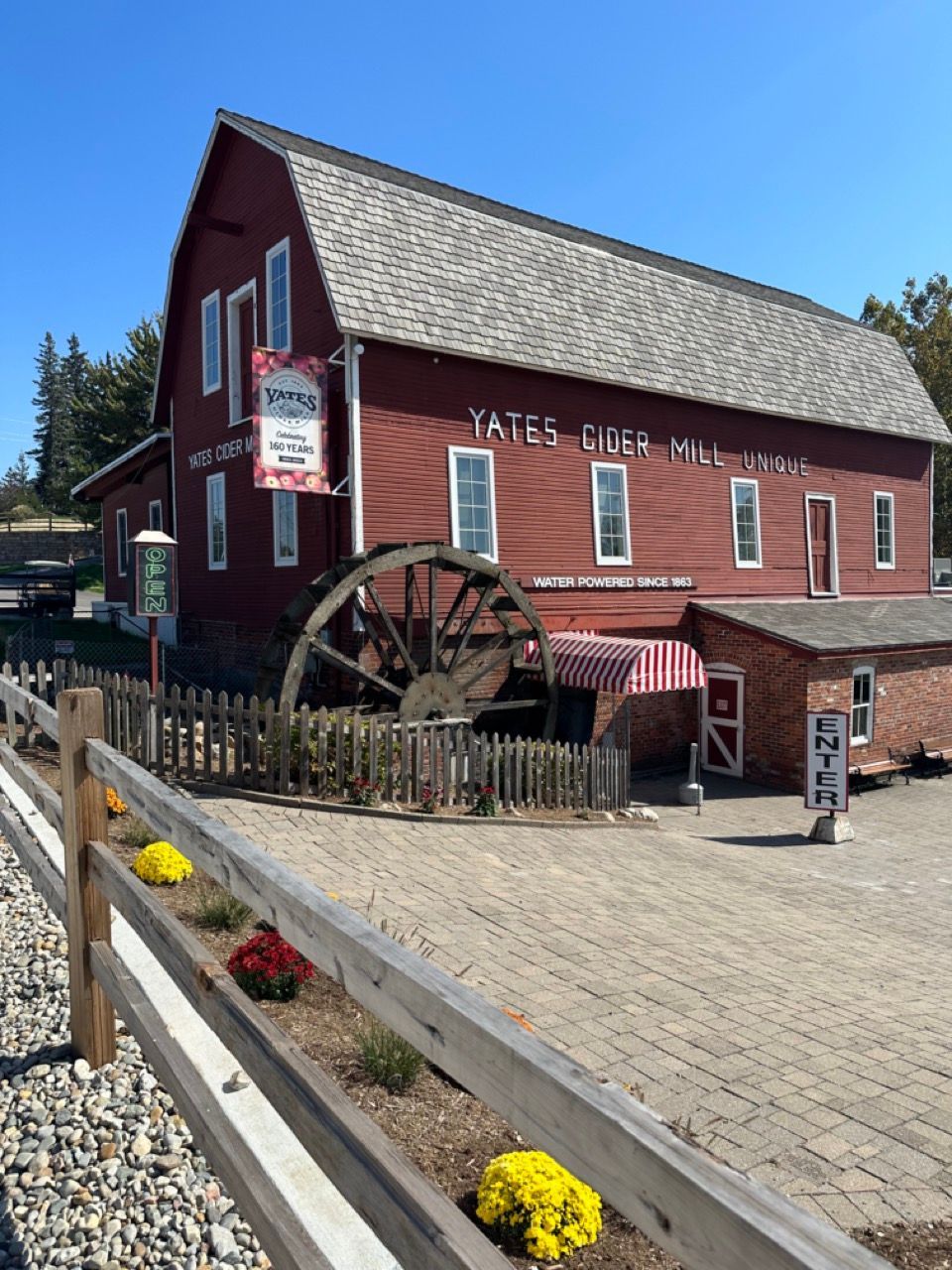 Red historic watermill building with large water wheel and flower-lined walkway under a clear blue sky