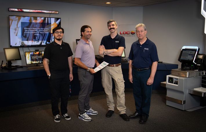 Four people standing in an office, smiling and holding a certificate beside a wall-mounted screen.