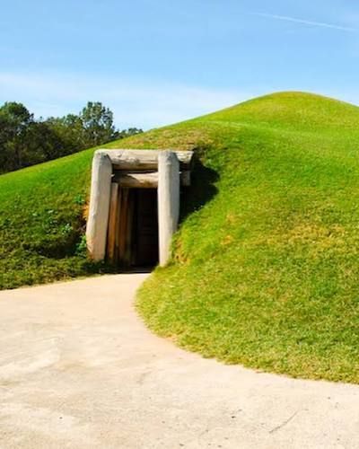 Grassy hillside with a small tunnel entrance beside a paved path under a blue sky