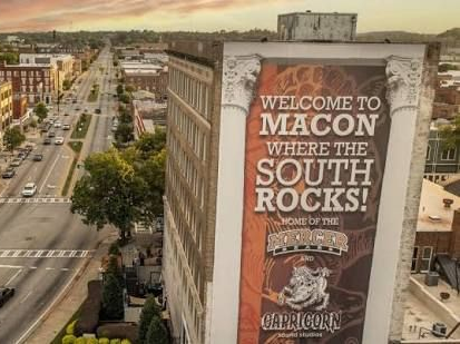 Macon billboard reading “WELCOME TO MACON WHERE THE SOUTH ROCKS!” beside a city street at sunset