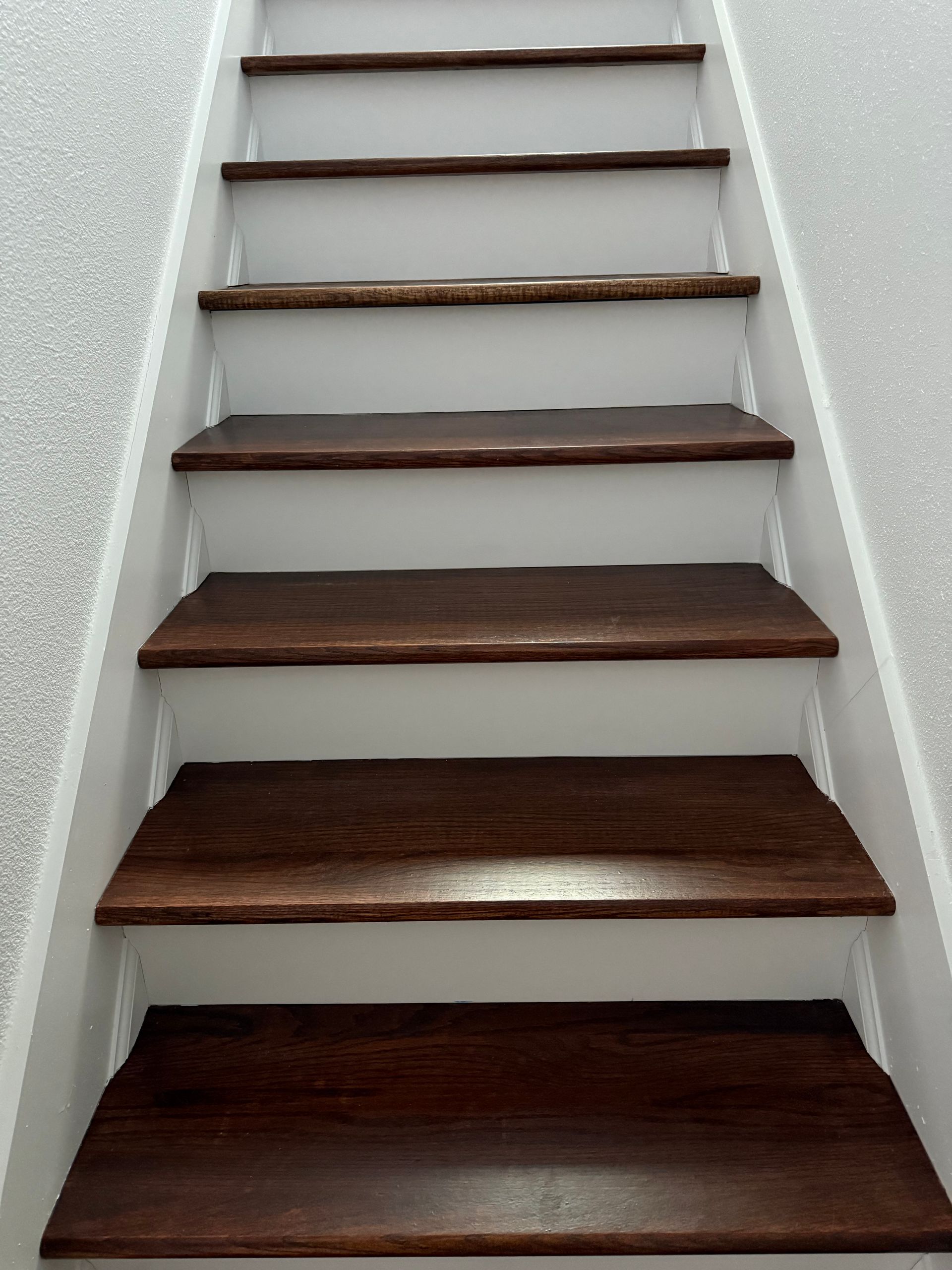 Dark wood stairs with white risers and brackets, leading upwards against a textured, light gray wall.