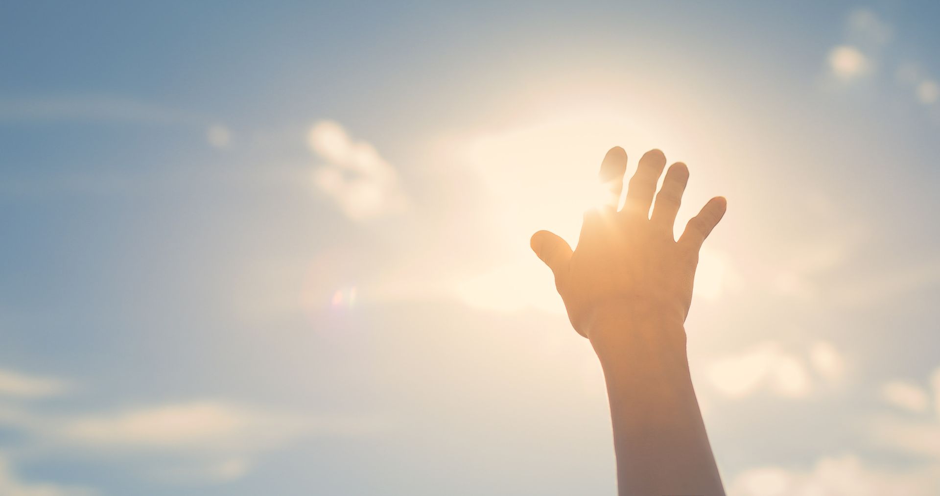Hand reaching up towards bright sun in a blue sky.
