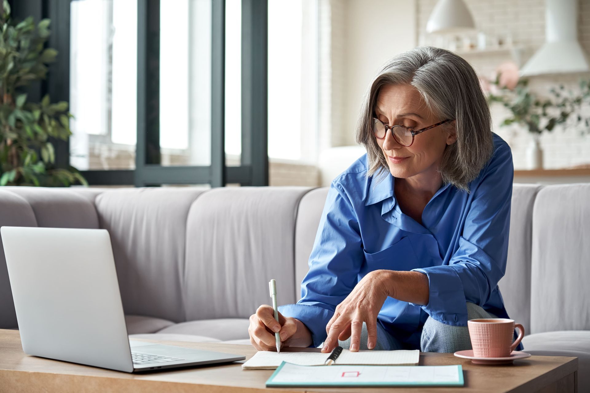 Woman with glasses, gray hair, writing on a notepad while using a laptop on a sofa.