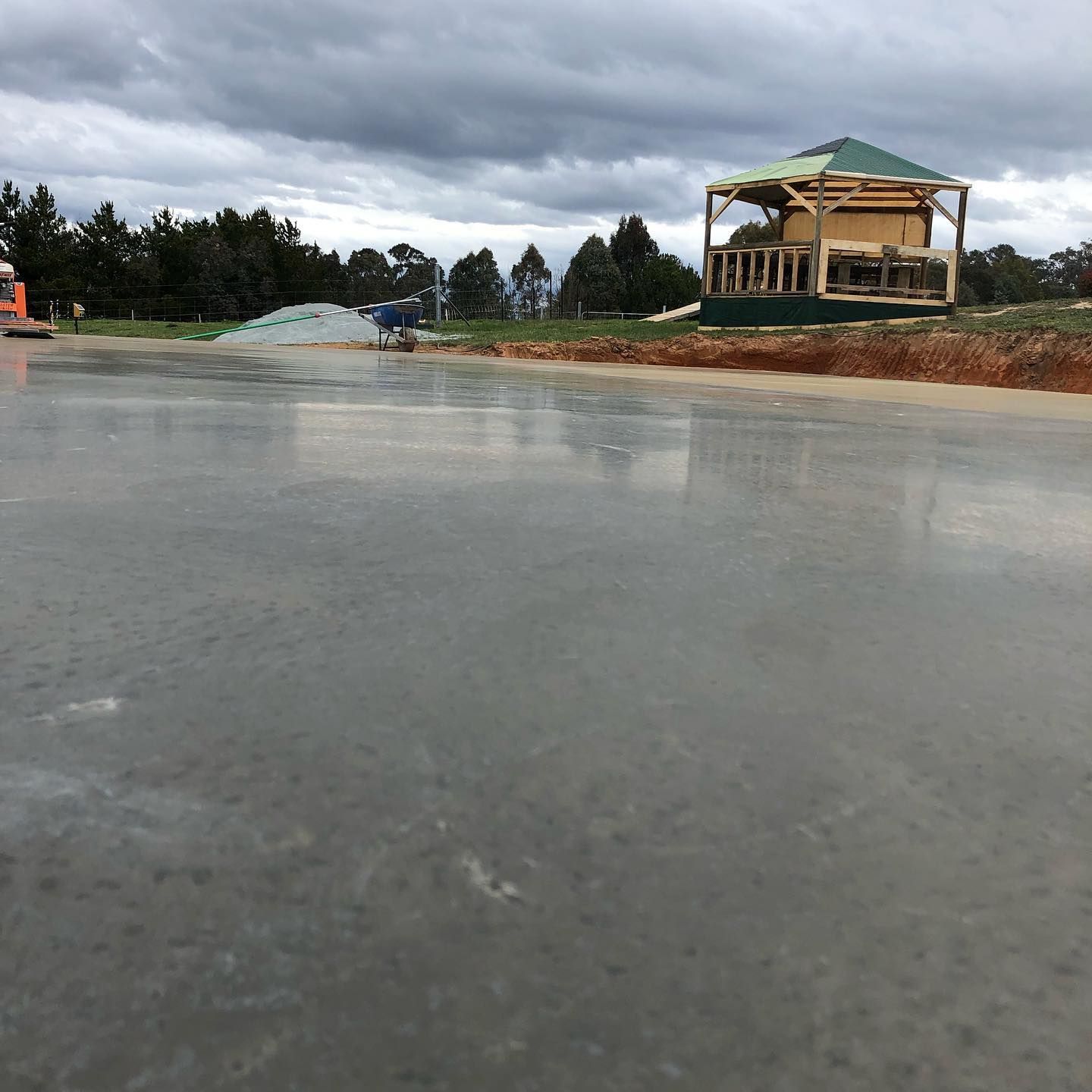 Wet concrete slab at a construction site with a partly built house in the background under cloudy skies