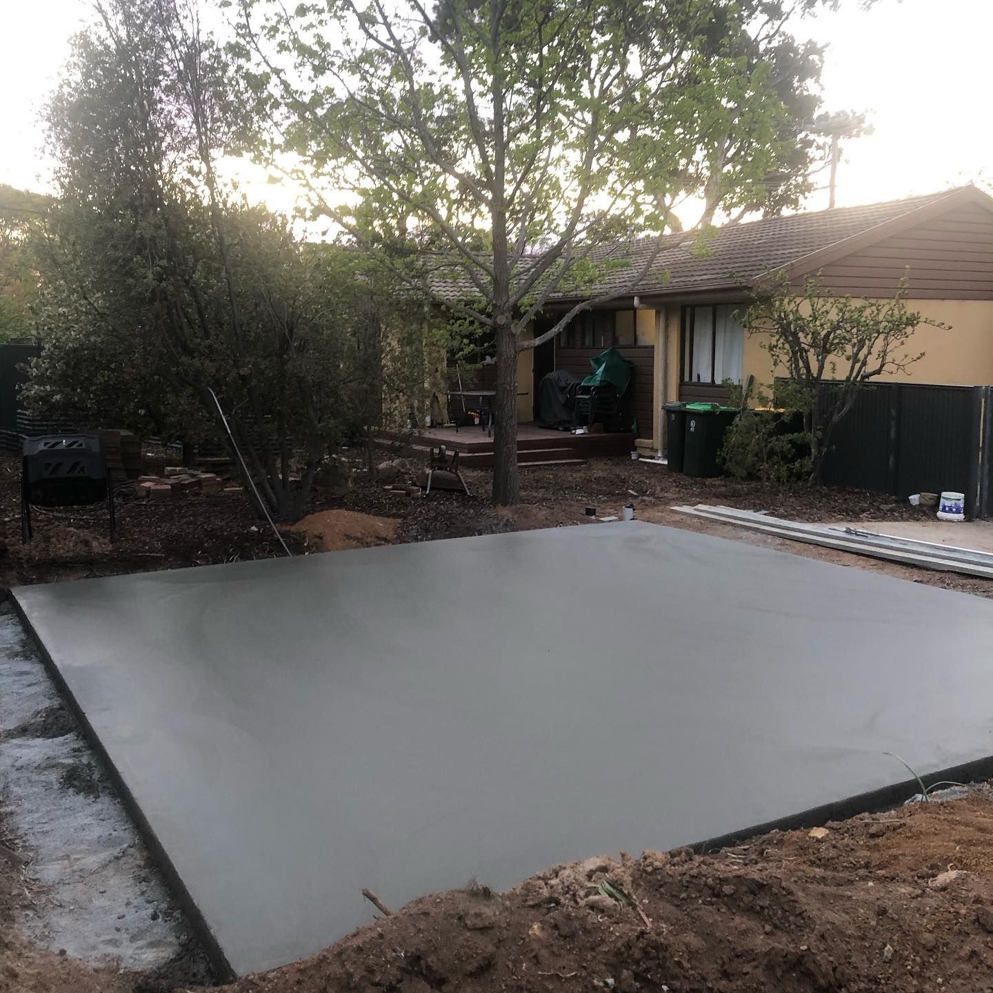 Freshly poured concrete slab in a backyard, bordered by dirt, trees, and a house at dusk