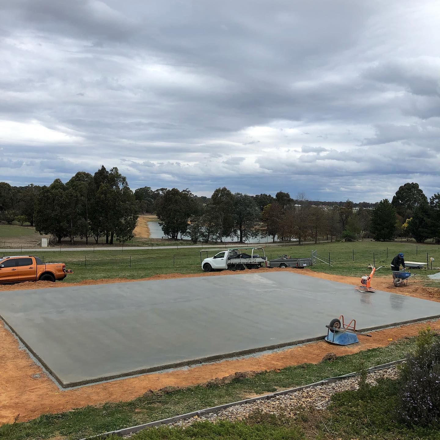 Construction site with a freshly poured concrete slab, workers, and parked vehicles under a cloudy sky