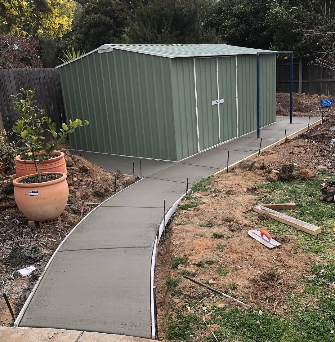 Green metal shed beside a curved concrete walkway in a backyard garden area