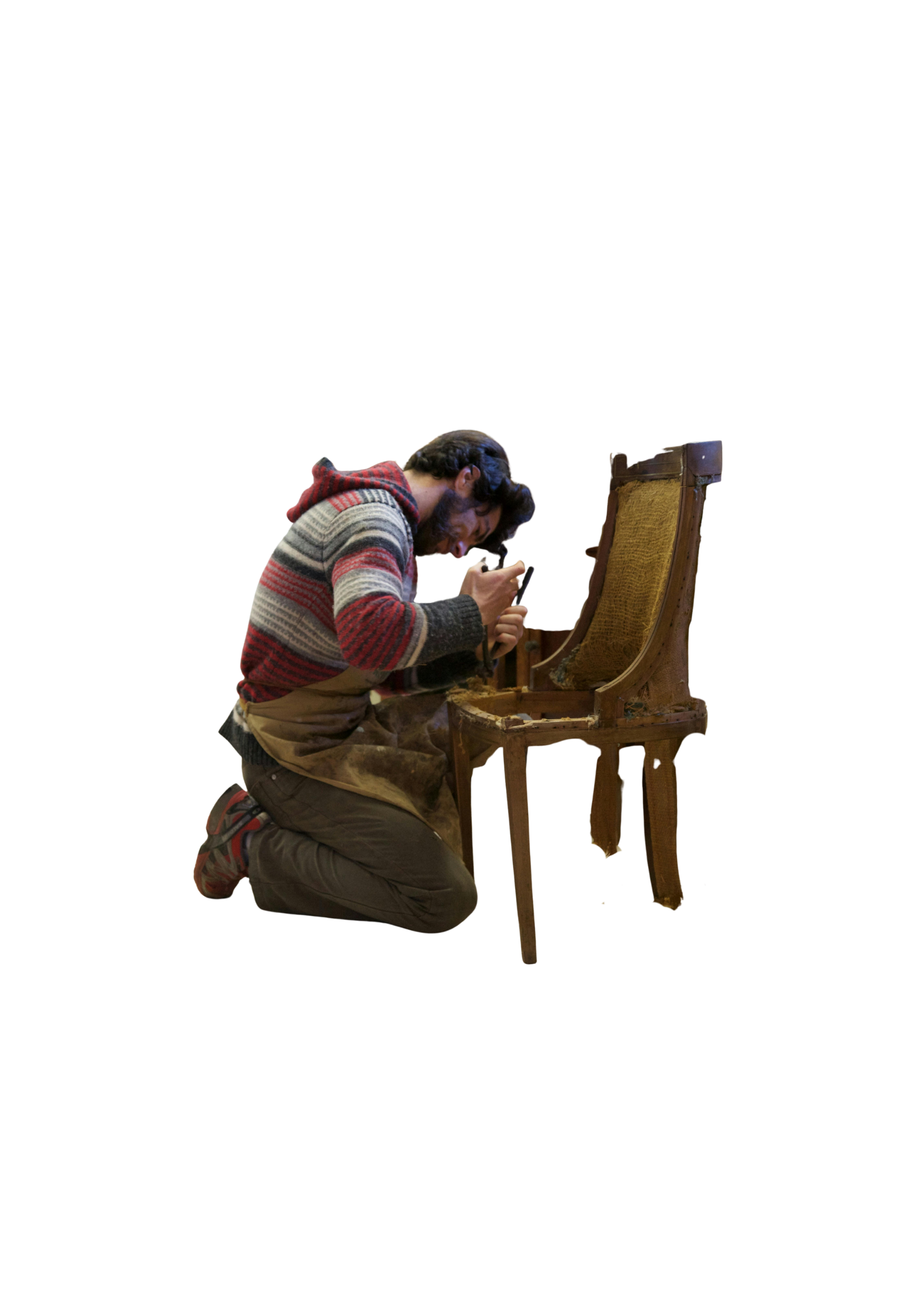 A man kneels, working on a wooden chair. He wears a red and white striped sweater.