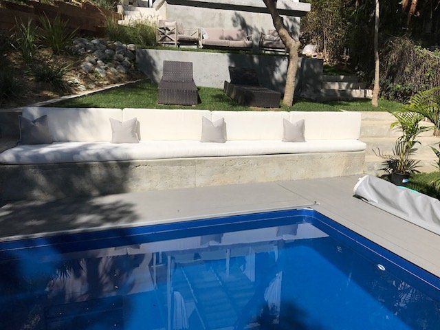 A concrete bench with cushions and pillows near a pool; in a backyard setting.
