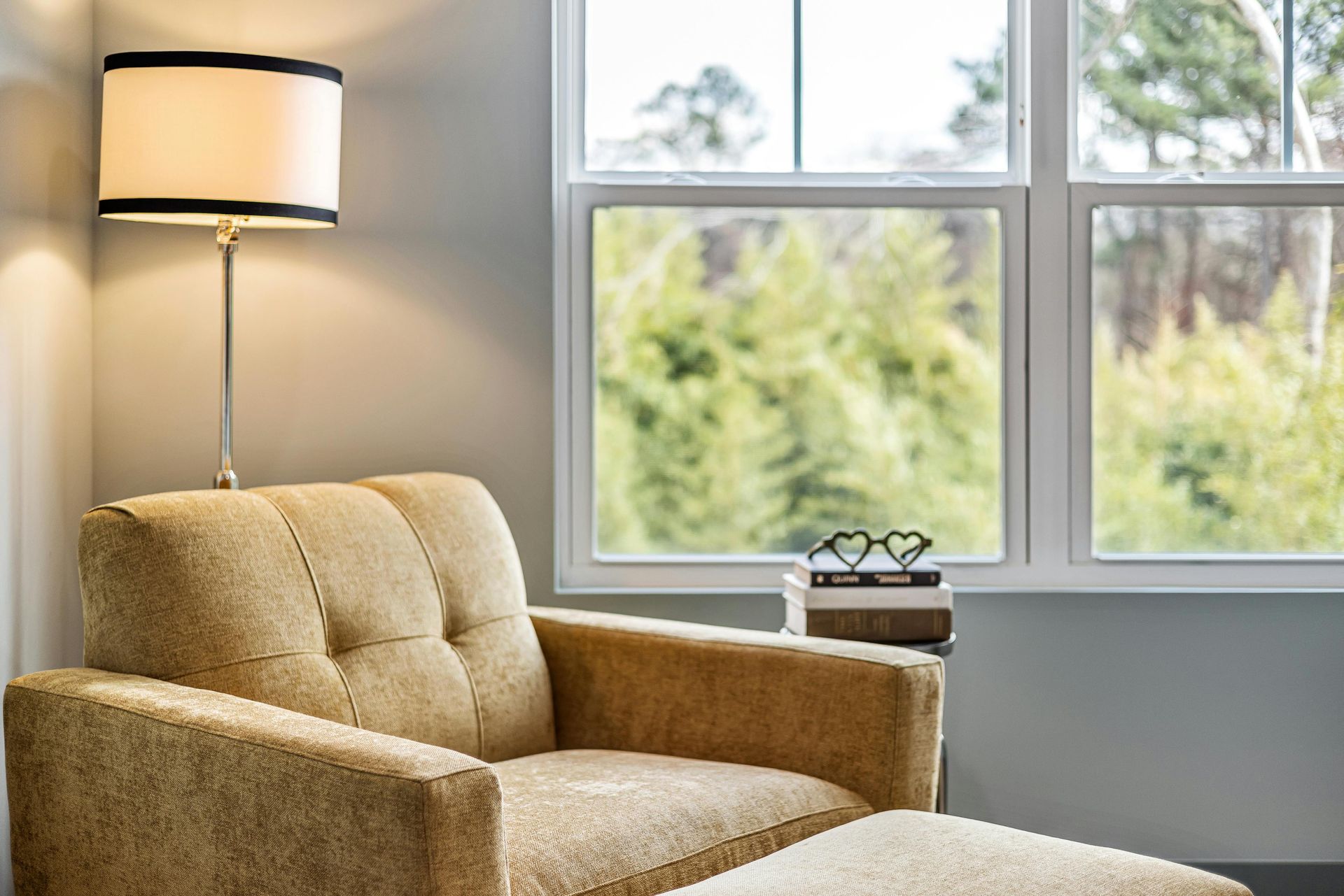 Beige armchair with a matching footstool next to a window overlooking trees. A lamp is next to the chair.