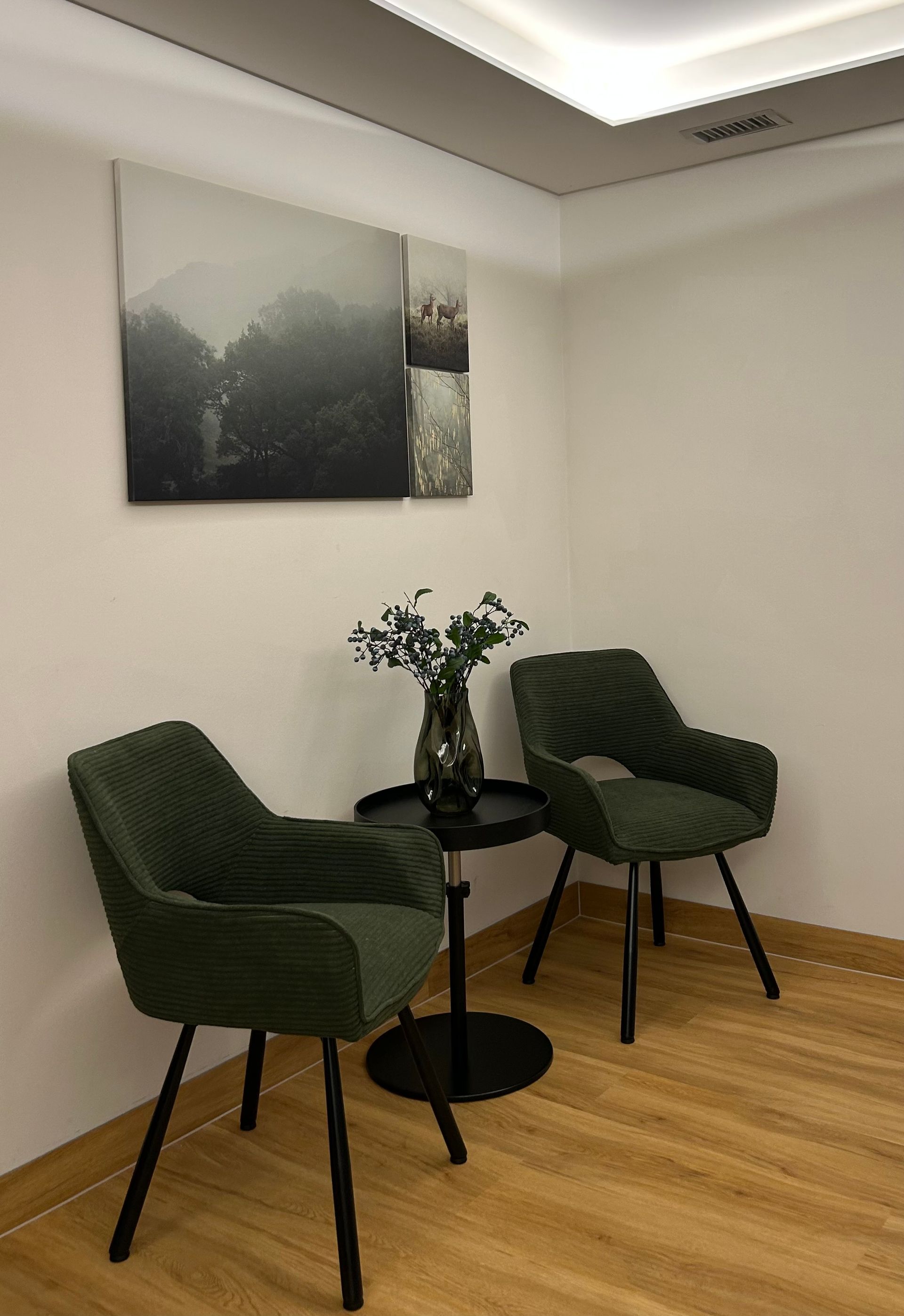 Small waiting area with two green chairs, a round black table, wall art, and wood flooring.