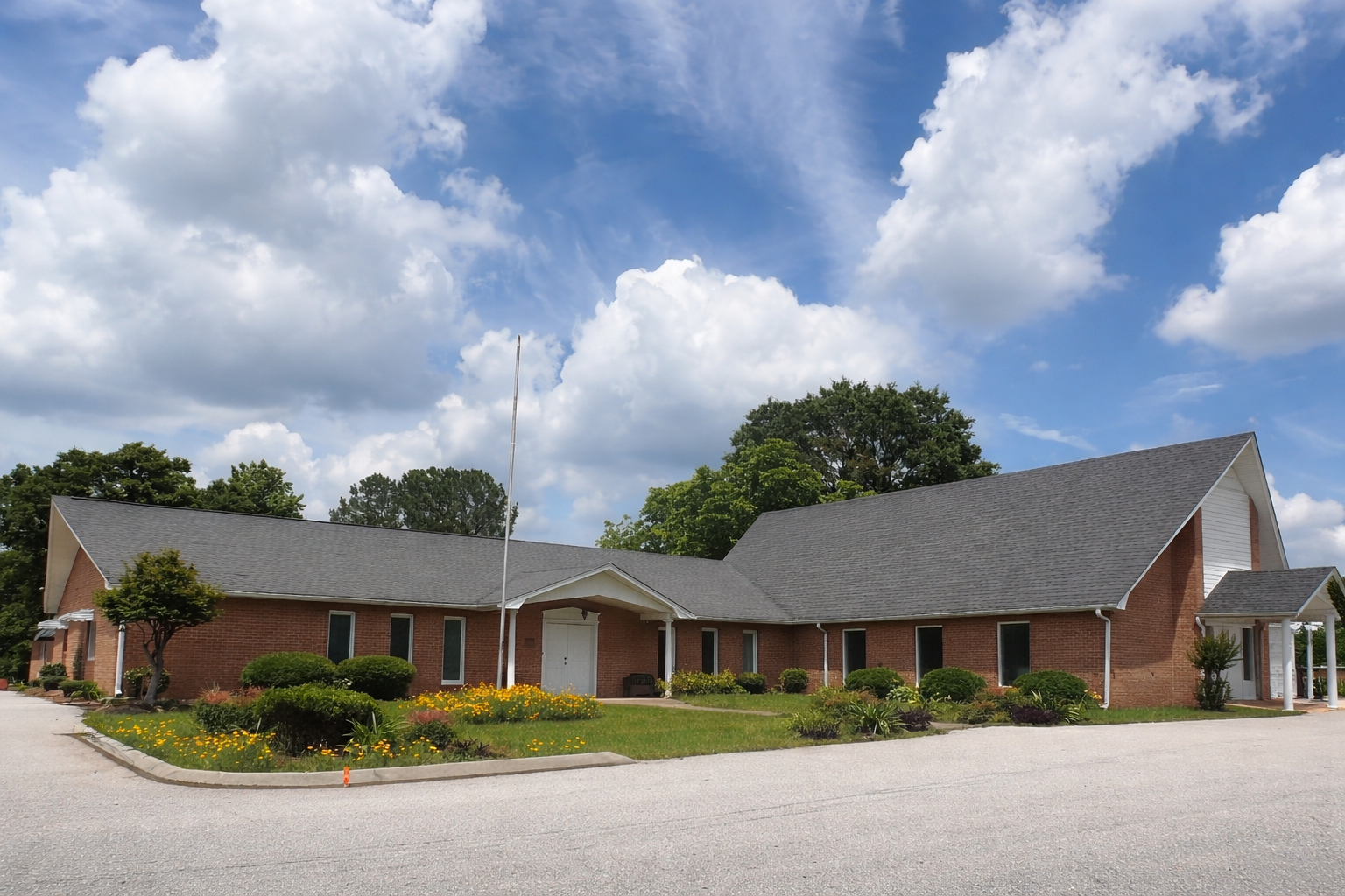 A single-story red brick church building with a gray roof and a gravel parking lot under a bright, cloudy blue sky.