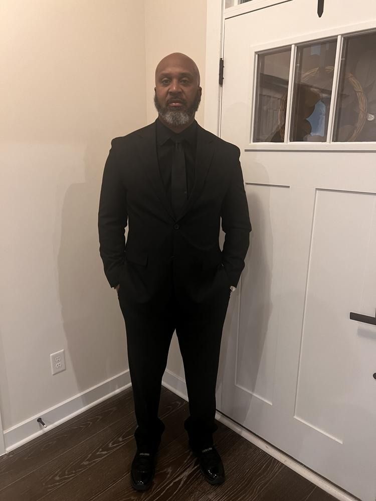 A person with a beard stands indoors near a white door, wearing a black suit and tie with hands in their pockets.