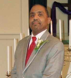 A person in a gray suit and red tie stands with a boutonniere, set against a backdrop of candles and draped fabric.