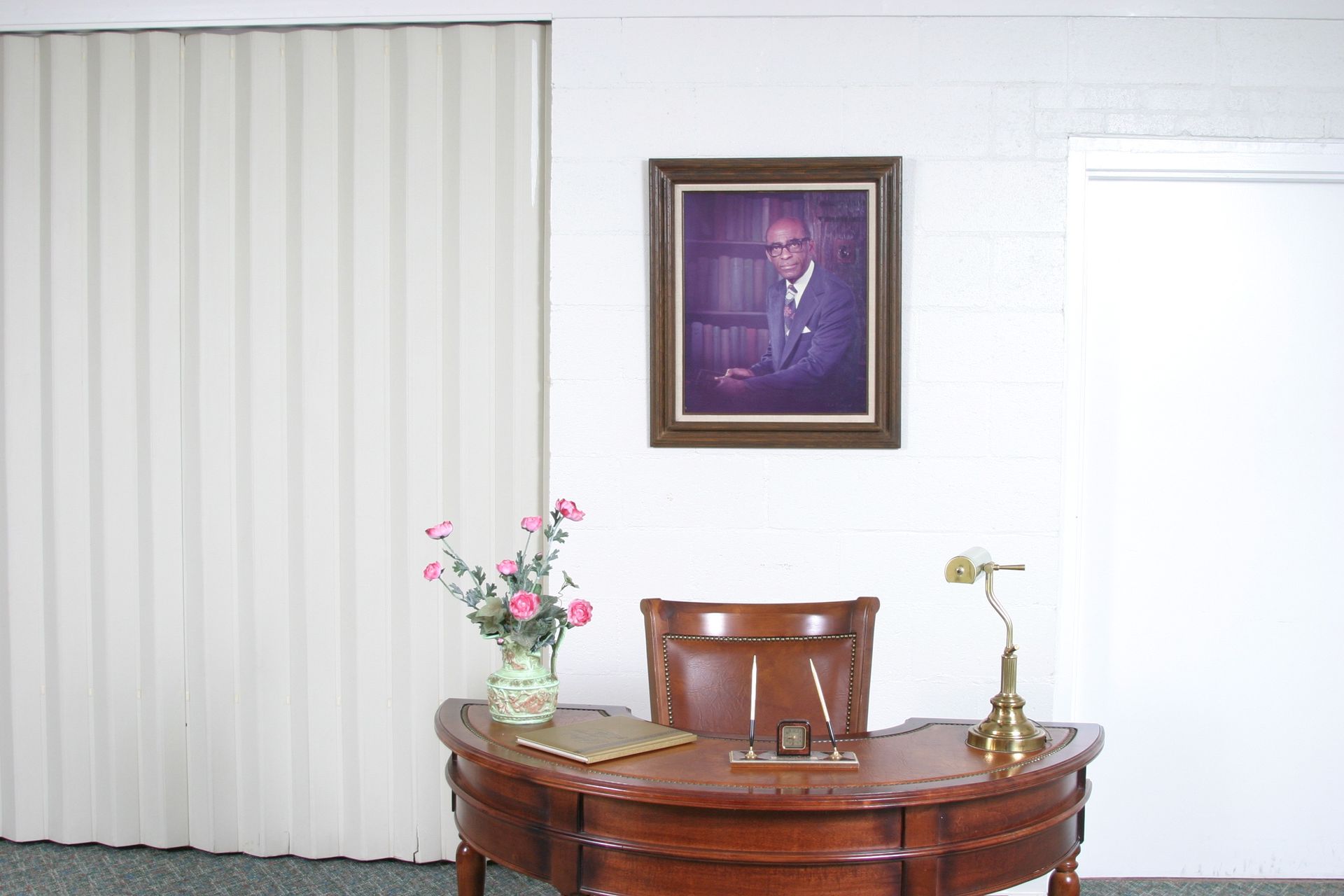 A wooden desk with a lamp, flowers, and pens stands before a wall featuring a framed portrait and vertical blinds.