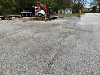 An excavator works at a paved construction site, loading debris into a dump truck parked near small buildings.