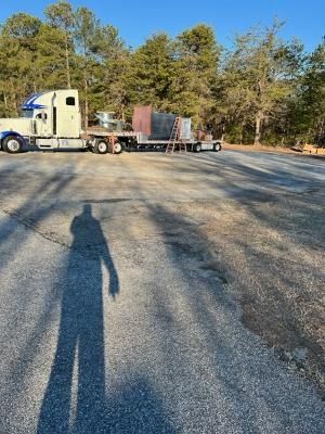 A semi-truck with a flatbed trailer hauling large metal industrial equipment parked on a gravel lot under a clear blue sky.