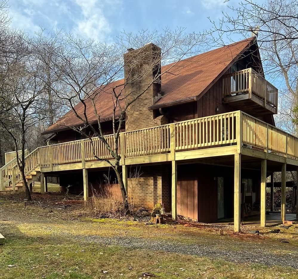 Wooden house with wraparound porch, stone chimney, and balcony in a wooded yard