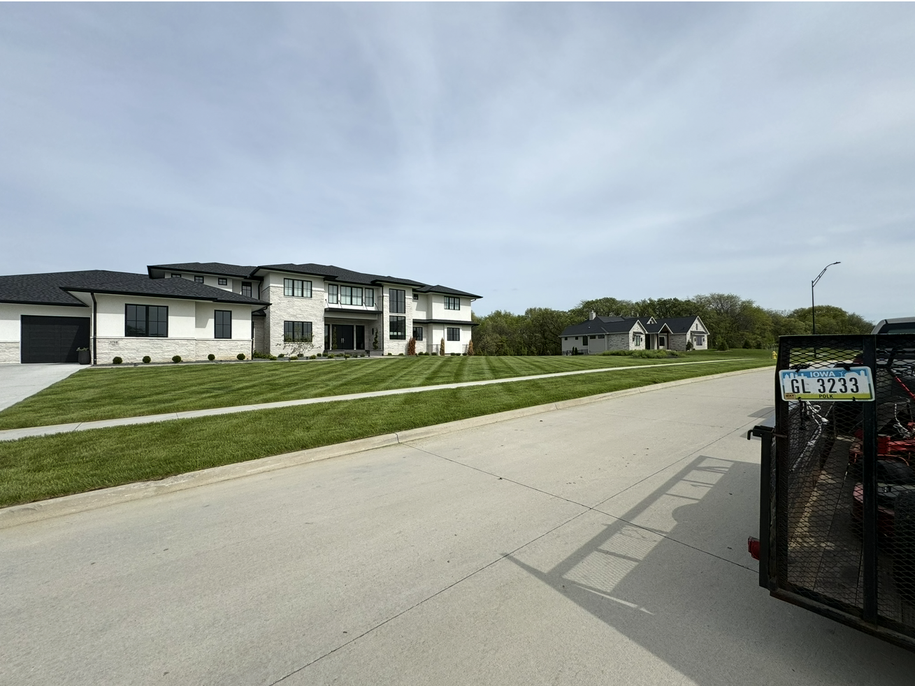 Quiet suburban street with modern white houses, grassy slopes, and a driveway gate on the right