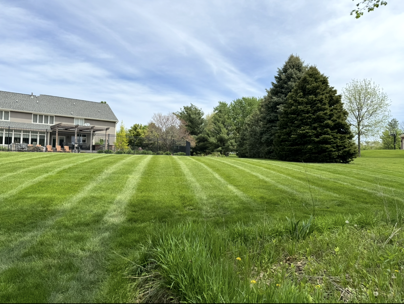Wide green lawn with striped grass, a house on the left, and trees under a partly cloudy sky.