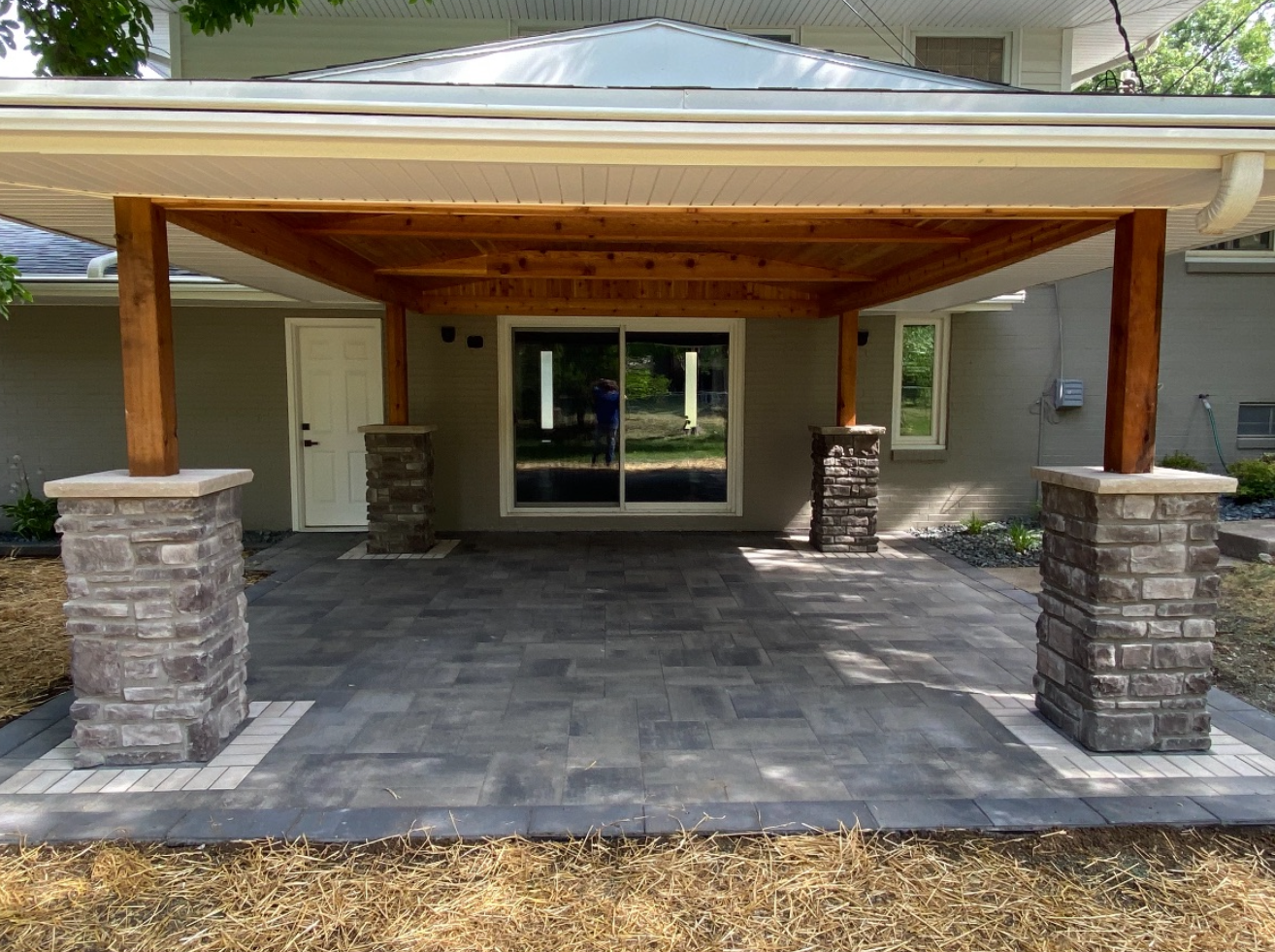 Covered stone patio with wooden roof and stone columns in front of a house entrance