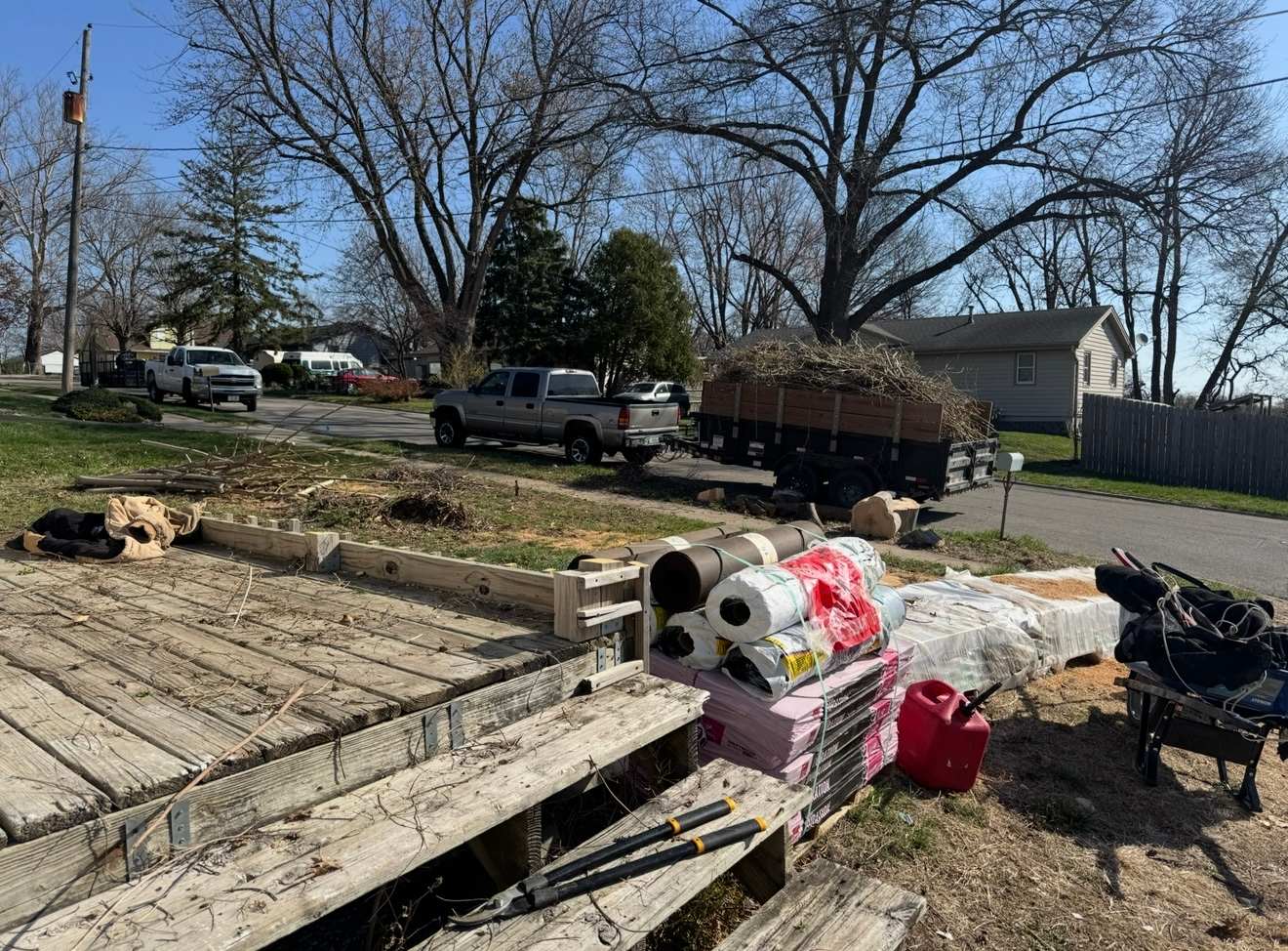 Yard with a truck, trailer, and piled trash, including a mattress and debris, beside a street and bare trees.