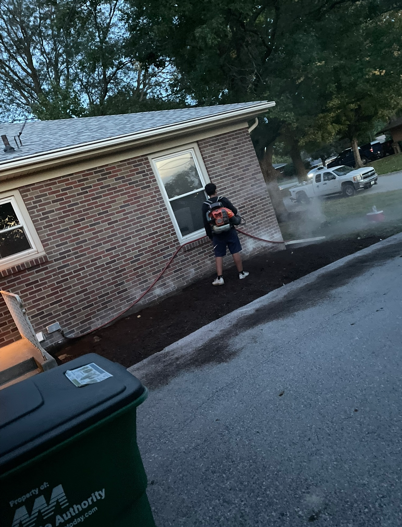 Person standing on a driveway beside a brick house, near a green trash bin, with trees in the background.