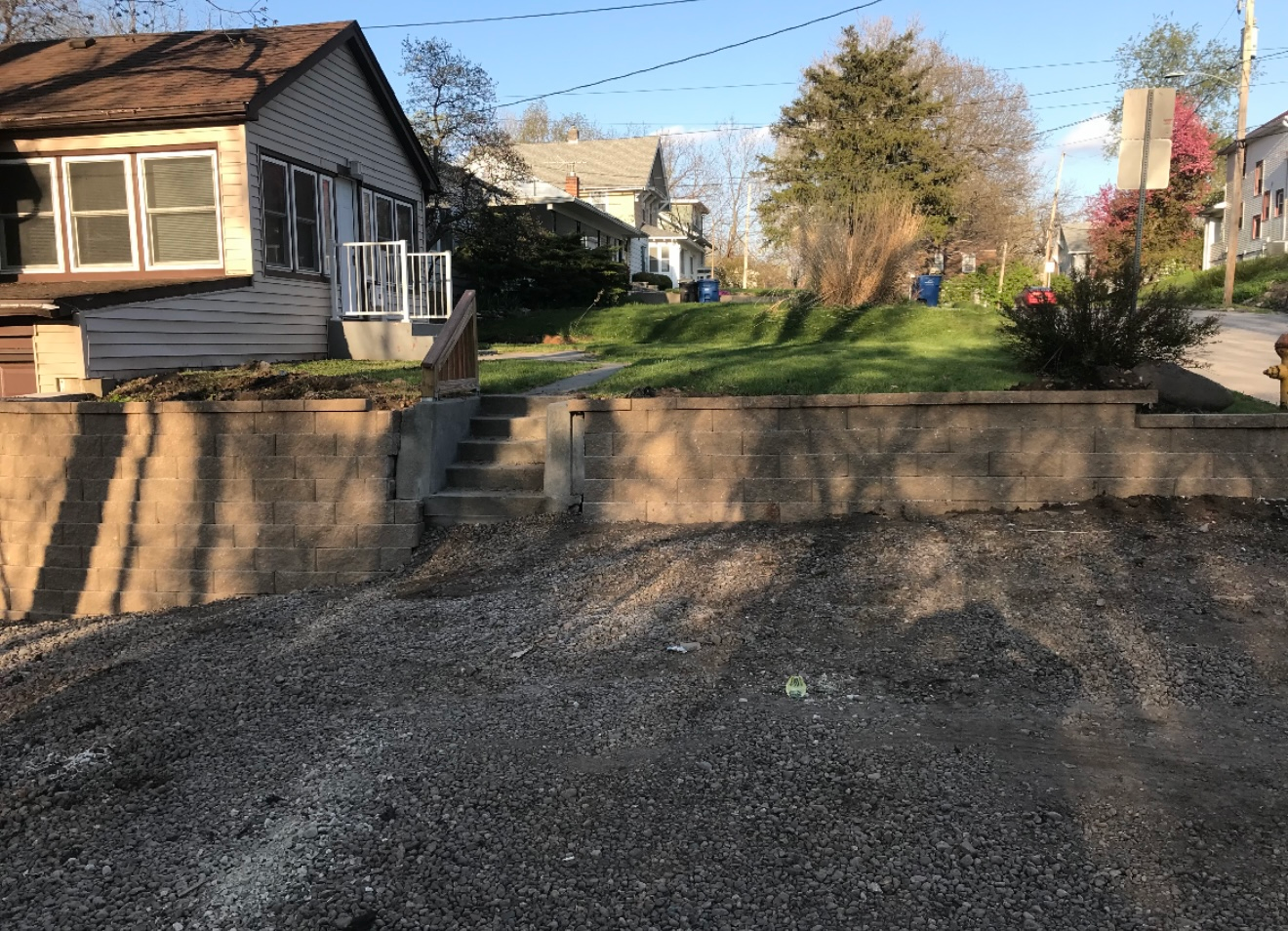 Residential yard with gravel driveway, low retaining wall, and steps leading to a house on a sunny day