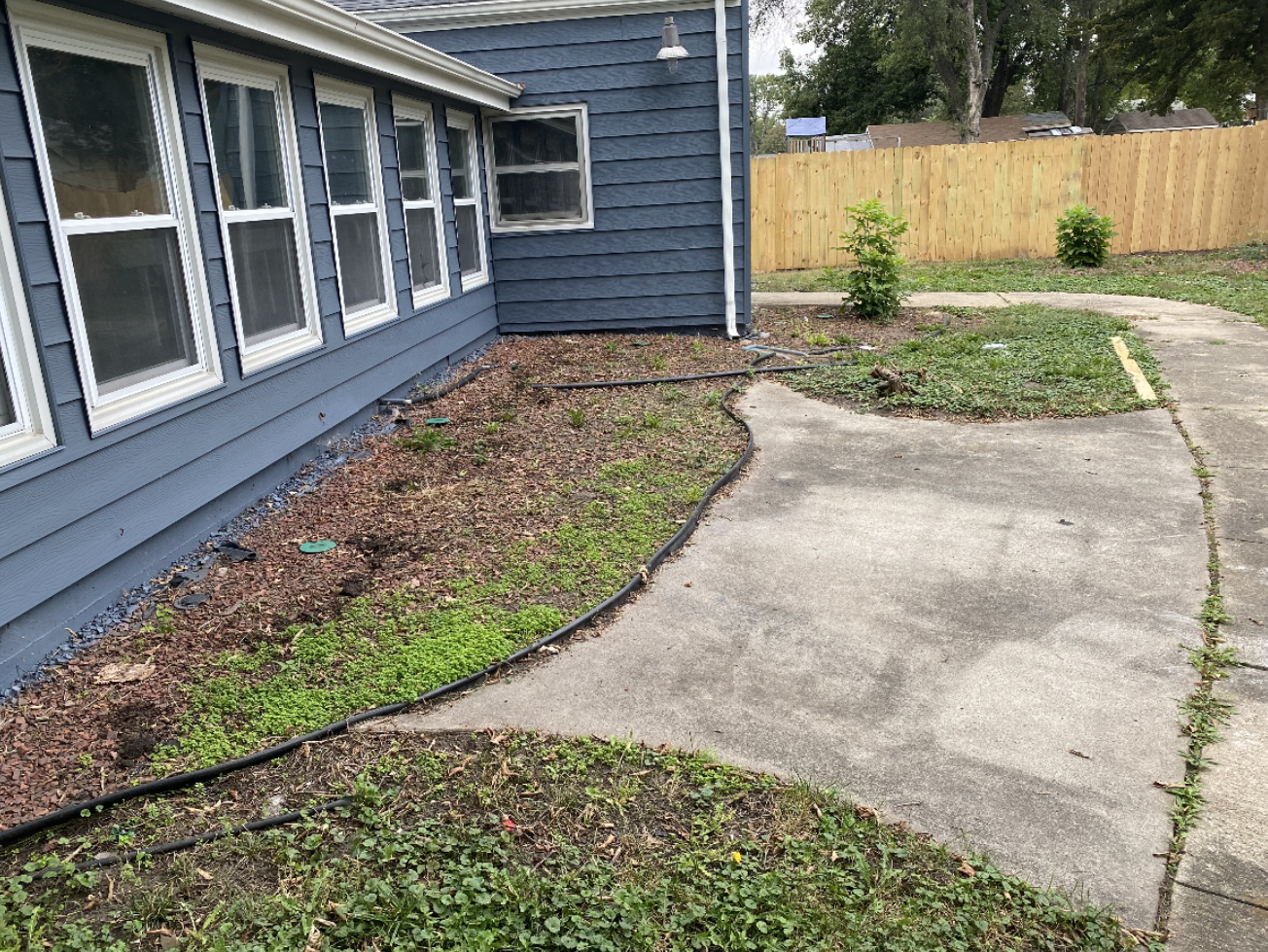 Curved concrete walkway beside a blue house, with mulch beds and a fenced yard.