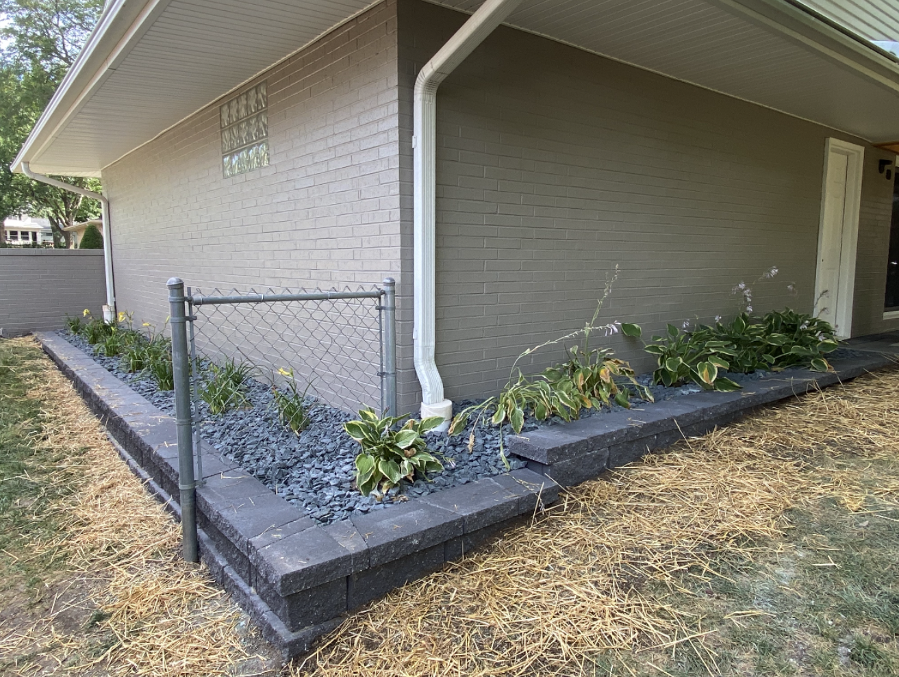 Small yard corner with a chain-link fence, raised planter bed, and a beige house wall under an overhang