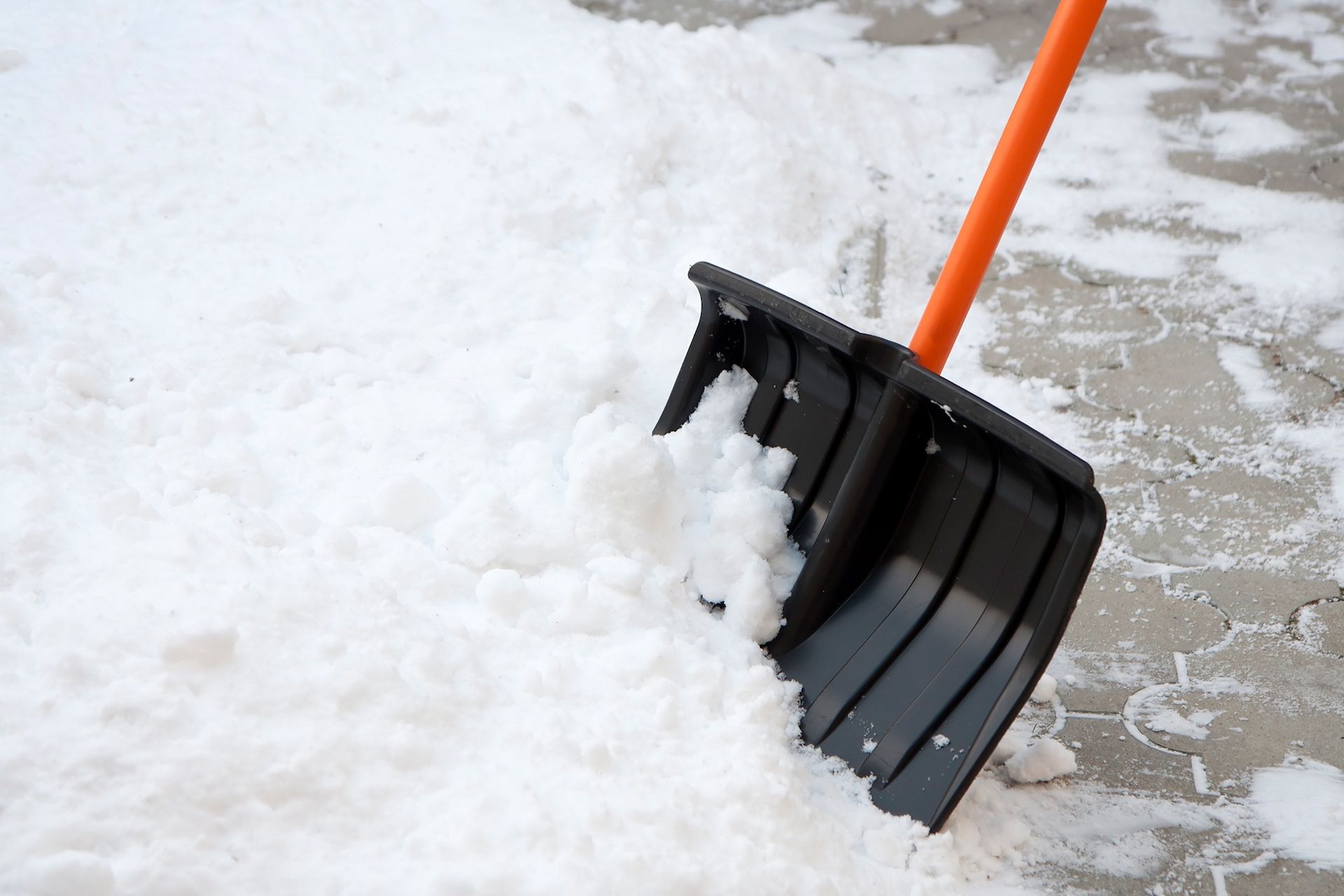 A black snow shovel with an orange handle clears a path through fresh snow on a paved surface.