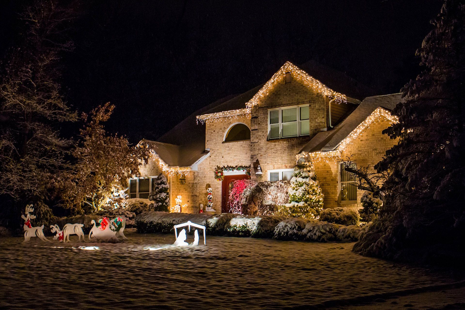 A two-story brick house at night decorated with Christmas lights, with a nativity scene and reindeer on the snowy lawn.