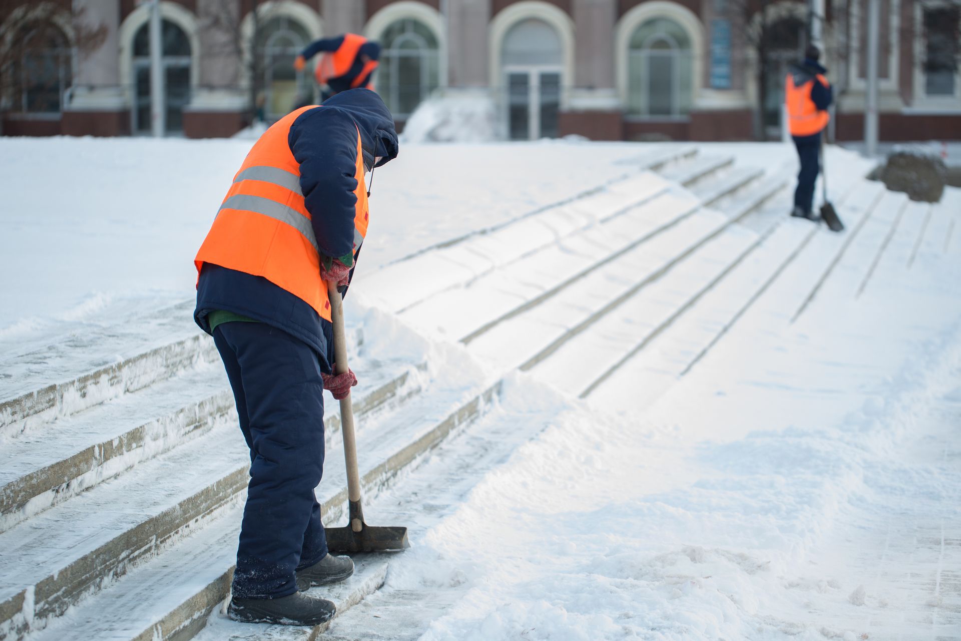 Workers in orange vests clearing snow from outdoor steps with shovels