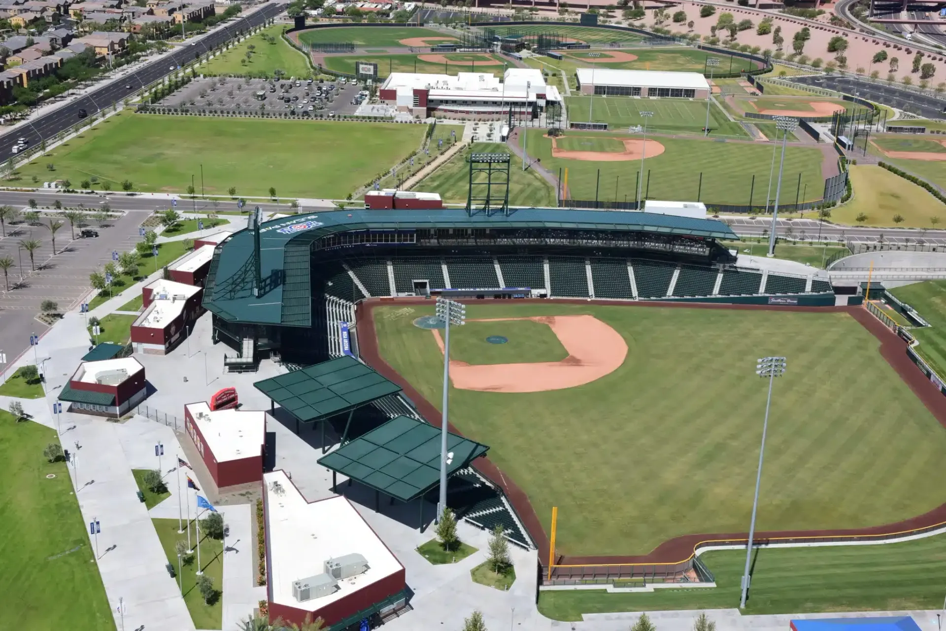 Aerial view of a baseball stadium with multiple fields and buildings; green grass, brown dirt, and blue sky.