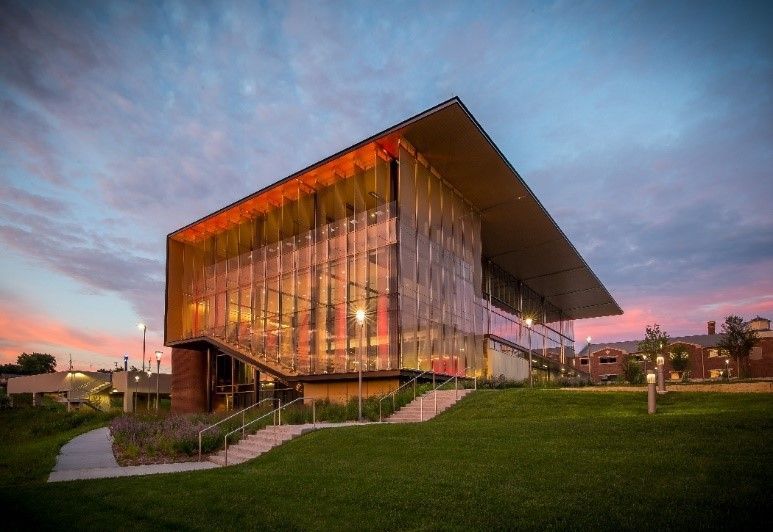 Modern building with glass facade, cantilevered roof, and orange and gold lighting, set against a sunset sky.