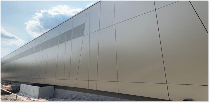 Exterior of a modern building with silver metal panels and a ventilation strip along the roofline. Blue sky with clouds.