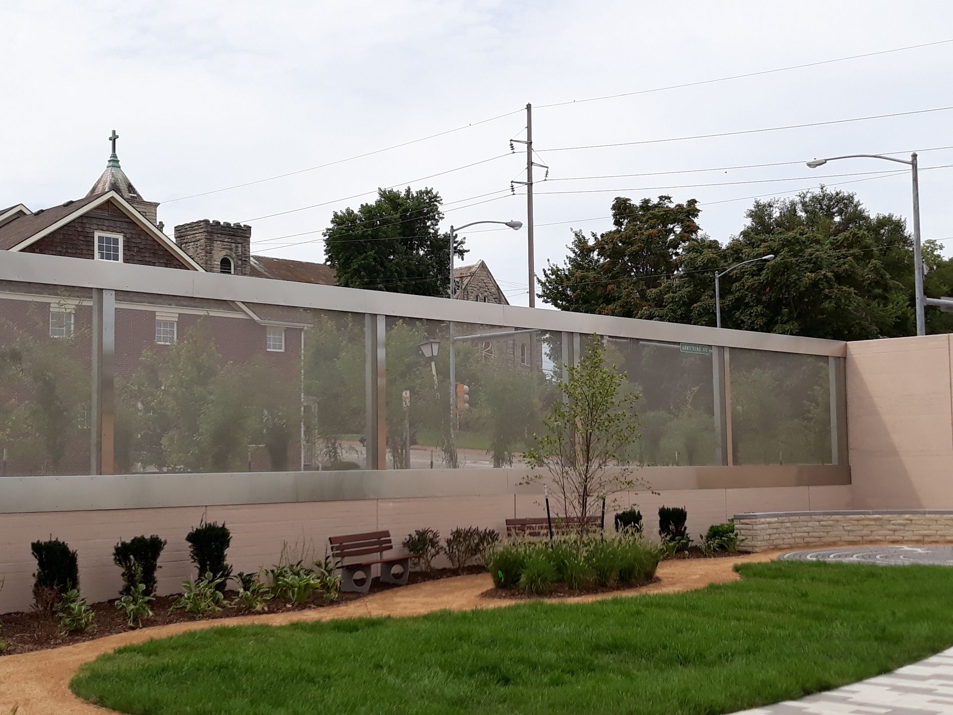 A view of a garden with a mesh screen wall. Benches and path in the foreground, church in the background.