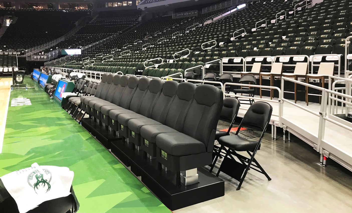 Basketball arena bench, black seats, green court, empty green seats in the background, Milwaukee Bucks logo visible.