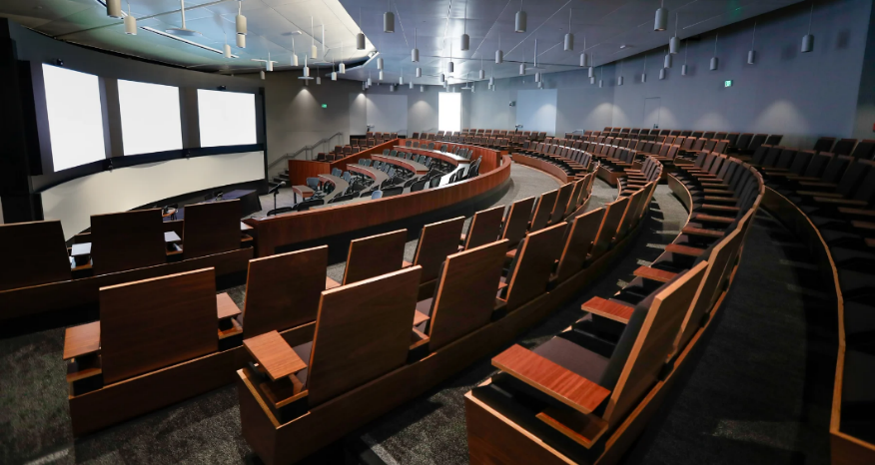 Curved lecture hall with rows of wooden seats facing a stage with three screens.