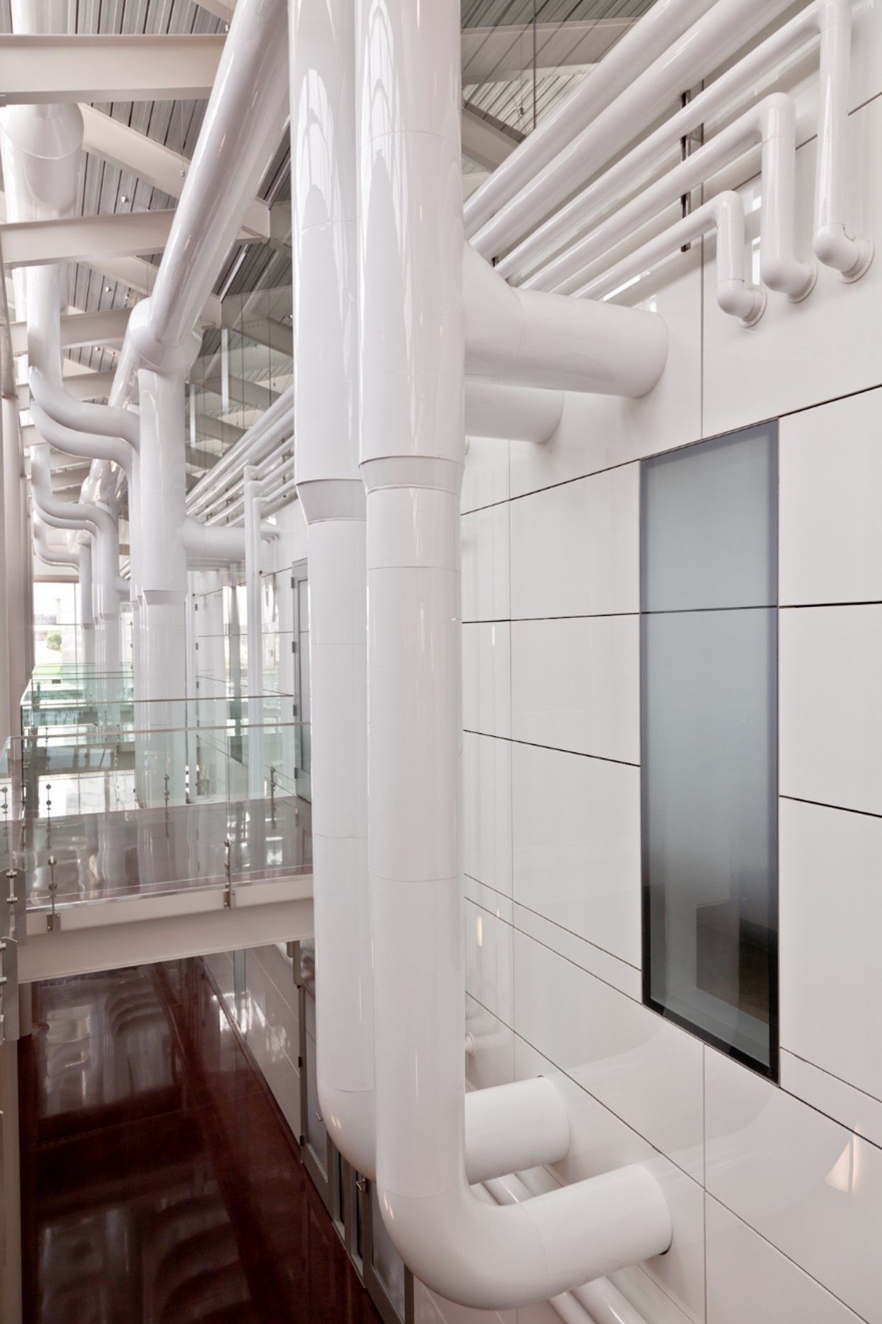 White pipes run along a white wall with a window. Balcony with glass railing and brown floor below.