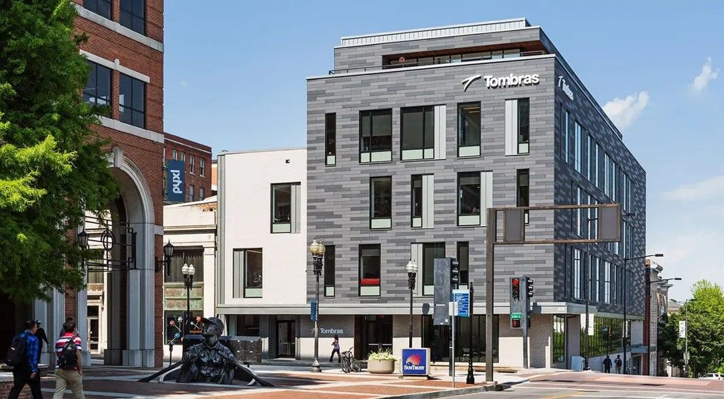 Modern multi-story building with textured gray facade, blue signage, and pedestrians on a sunny street.