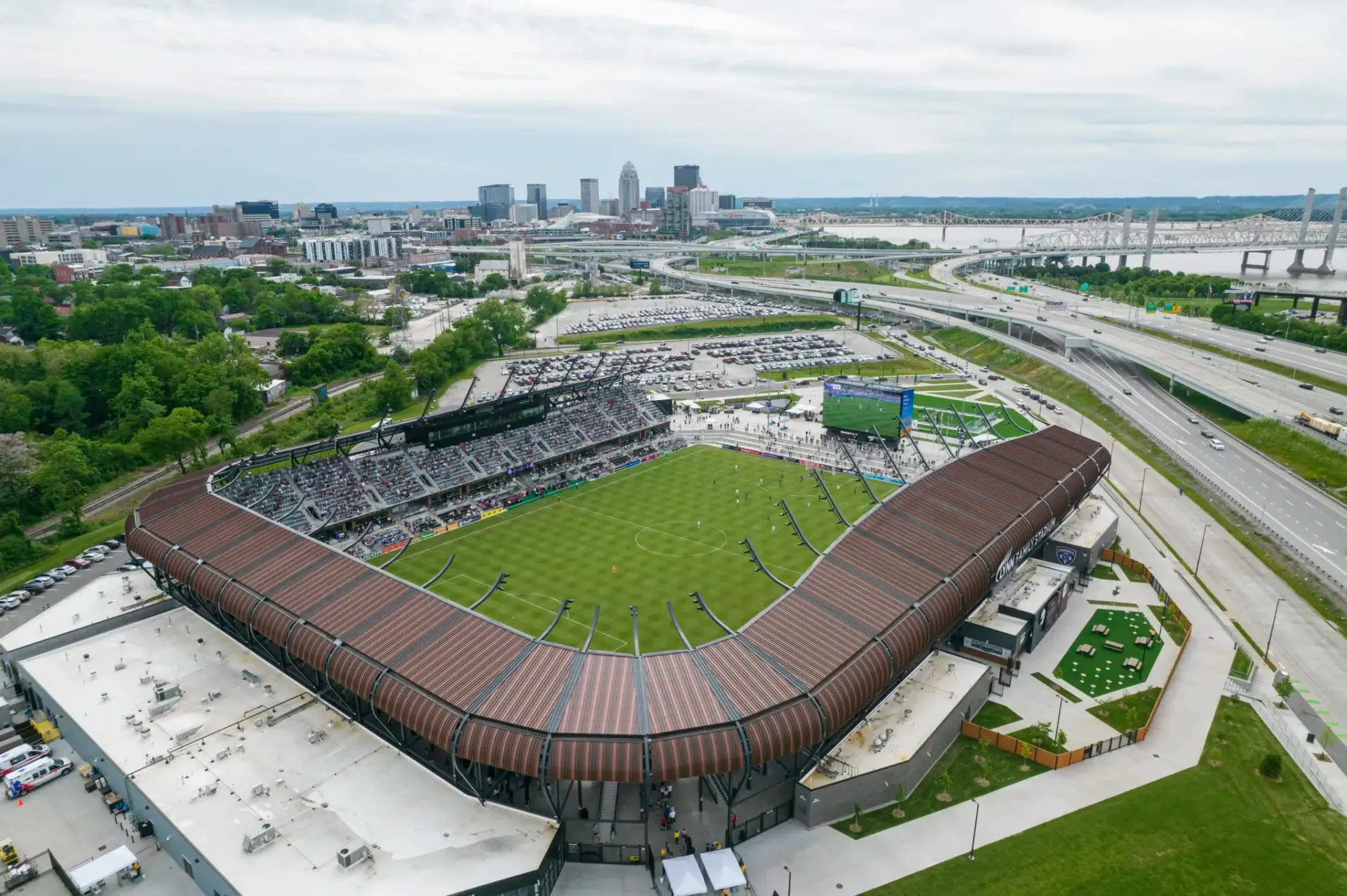 Aerial view of a soccer stadium with fans, green field, and city skyline in the background.