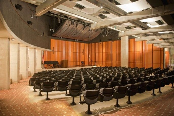 Auditorium interior with rows of black seats facing a stage with wooden paneling and a piano.