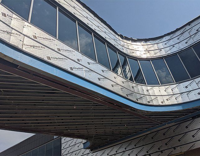 Curved building exterior under construction; exposed metal framework, reflective insulation, and windows against a blue sky.