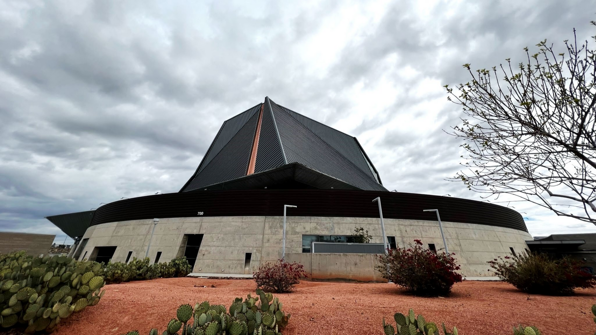 Modern, circular building with a dark, angular roof against a cloudy sky. Exterior view.
