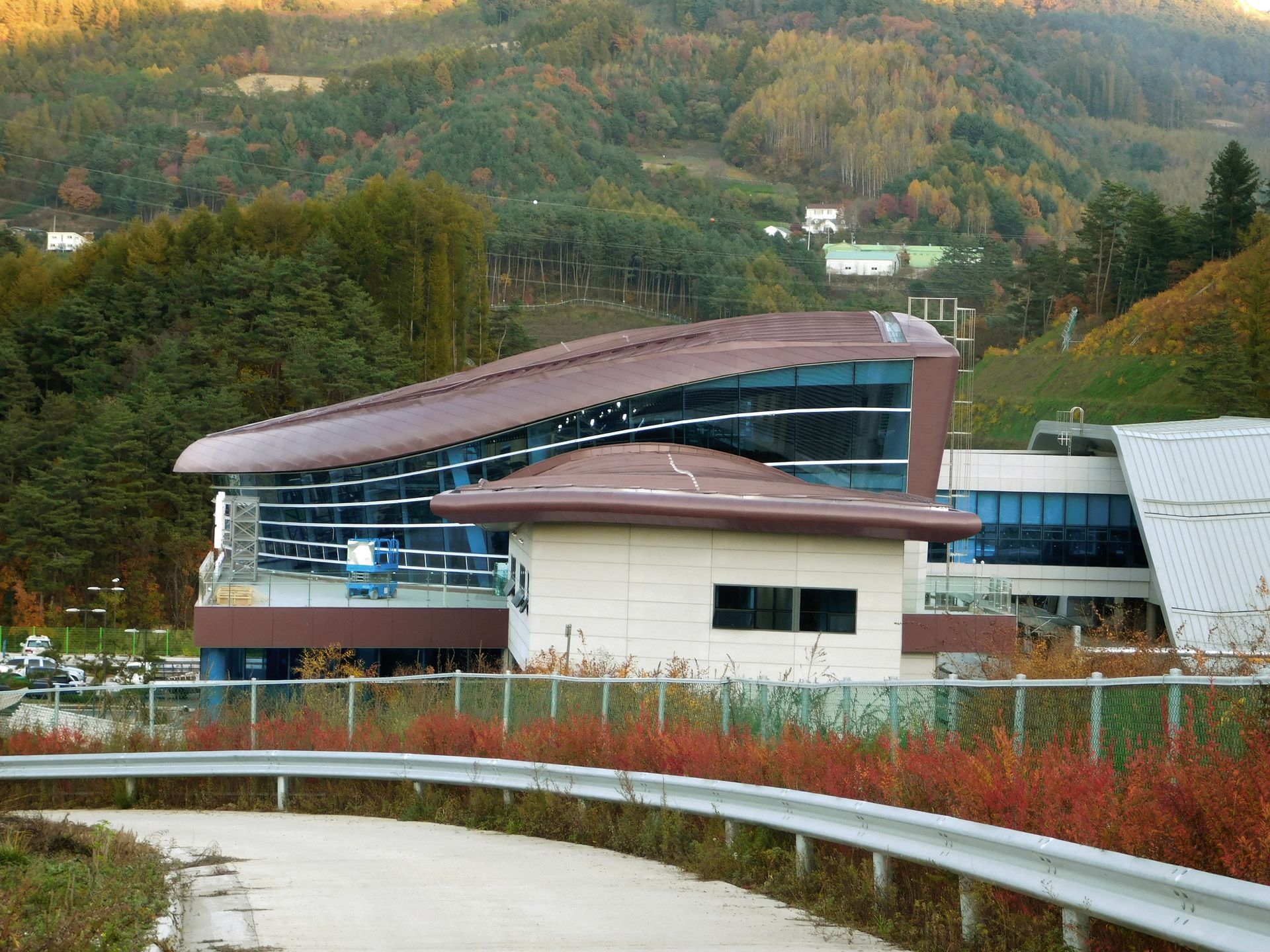 Modern building with a curved brown roof, glass facade, and a smaller attached structure, set against a forested hillside.