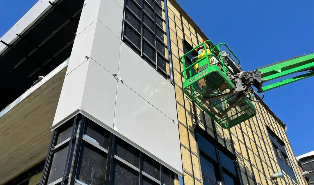 Construction worker in a lift installing white panels on a building exterior with large black-framed windows.